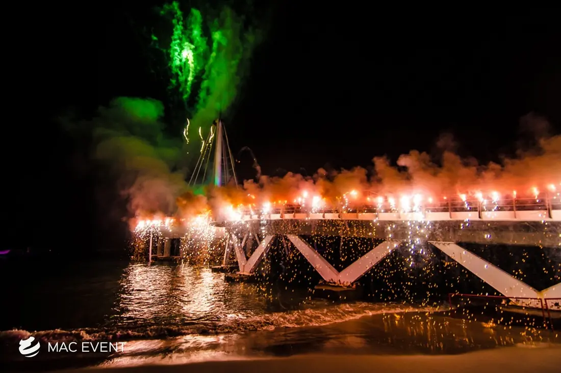 A spectacular fireworks display on a pier with bright green and orange lights reflecting on the water, creating a dramatic scene over the ocean.